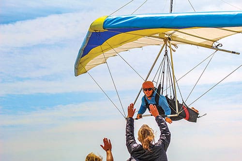 Celebrating 50 years in business, Kitty Hawk Kites has been “Teaching the World to Fly” since 1974. Pictured: Hang Gliding instructor Andy Thompson, being high fived by fellow instructors Charlie Trossbach and Emma Comer during the 51st Annual Hang Gliding Spectacular, back in May of 2023. Photo by Hannah Comer of Kitty Hawk Kites.
